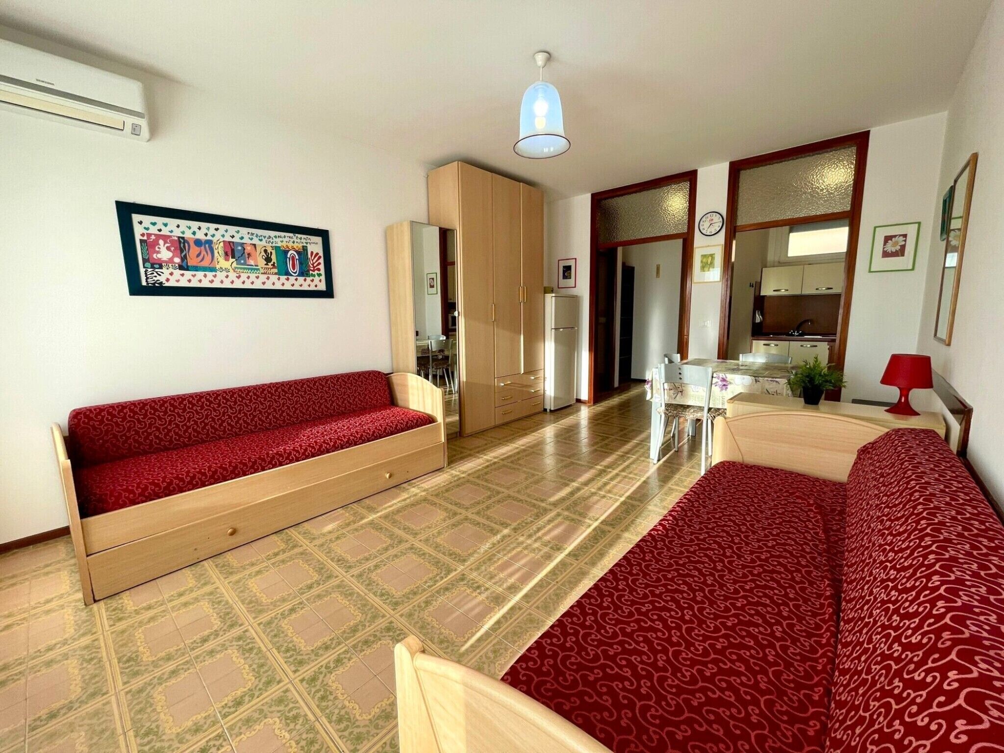 A living room with a red sofa, a wooden cabinet, a dining table with chairs, a kitchen area, and a clock on the wall.