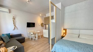 A modern, well-lit room with a grey sofa, a bed with a blue quilt, a dining table with chairs, and a TV mounted on the wall. - Studio Near the Beach-beahost (Caorle)