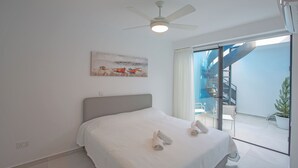 Un dormitorio con cama, ventilador de techo, un cuadro y vistas a un balcón con una silla y una planta en maceta.