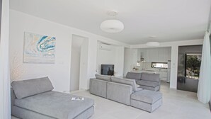 Una sala de estar moderna con sofás grises, un televisor de pantalla plana y una cocina al fondo.