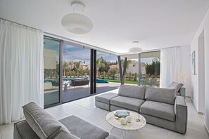 A modern living room with a grey sofa, a coffee table, and a view of a swimming pool and outdoor seating area. - Protaras Holiday Villa Cr3 (Paralimni)