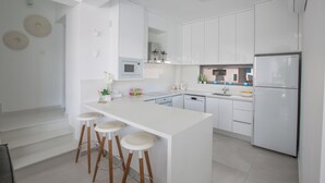 Una cocina moderna con armarios blancos, una isla central con taburetes, electrodomésticos de acero inoxidable y vista al área de estar.