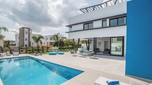 Un área moderna de piscina al aire libre con una piscina rectangular, sillas de descanso y un edificio de dos pisos con grandes ventanales.