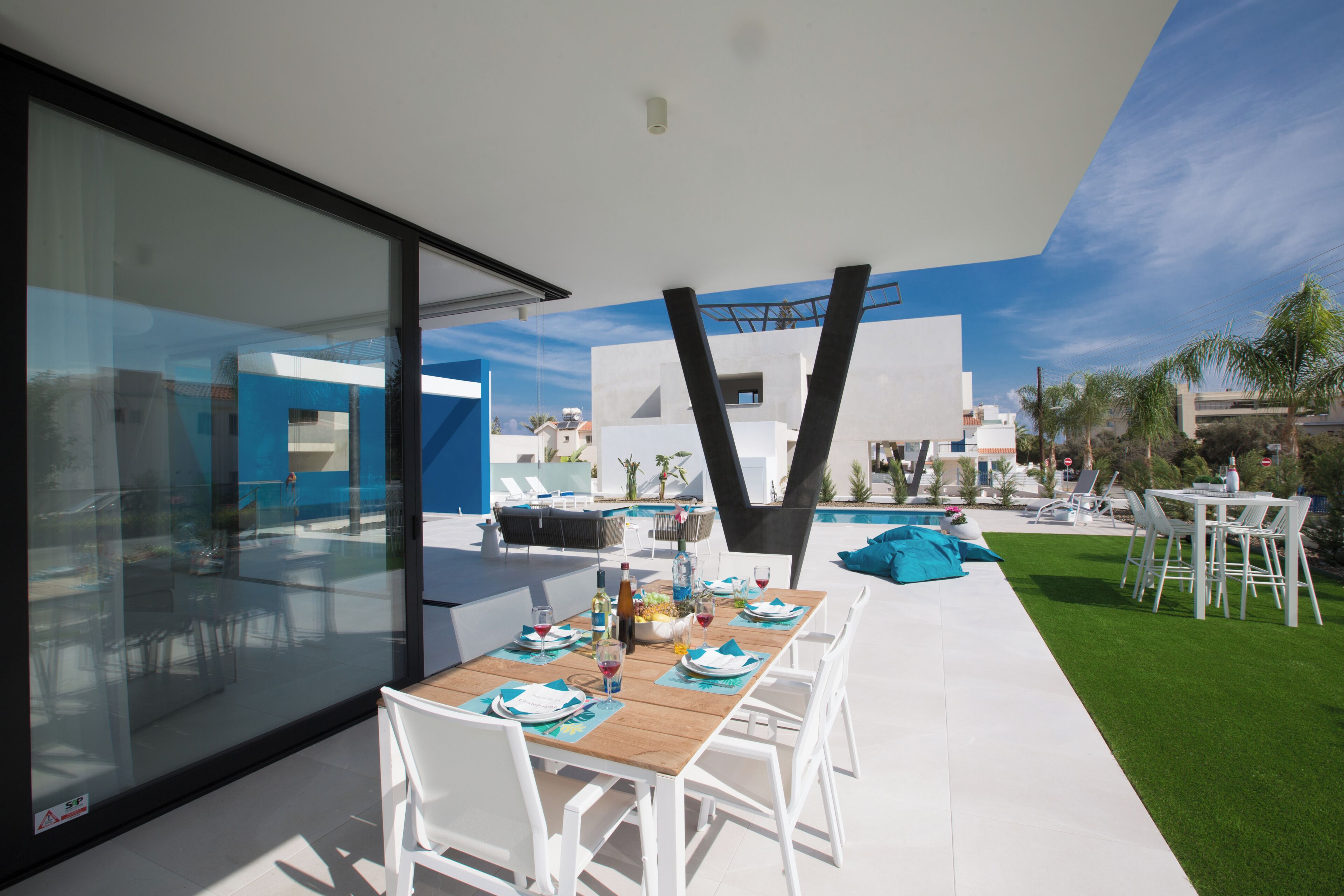 Un área de comedor al aire libre con una mesa larga dispuesta para seis personas, rodeada de sillas blancas, con una piscina y follaje al fondo.