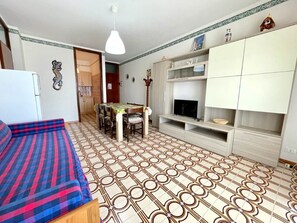 A compact living space with a sofa, dining table, TV stand, and patterned floor tiles. - Comfy Flat With Terrace 250mt From the sea (Caorle)