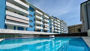 En swimmingpool med fliseterrasse, omgivet af en bygning med flere etager og altaner.