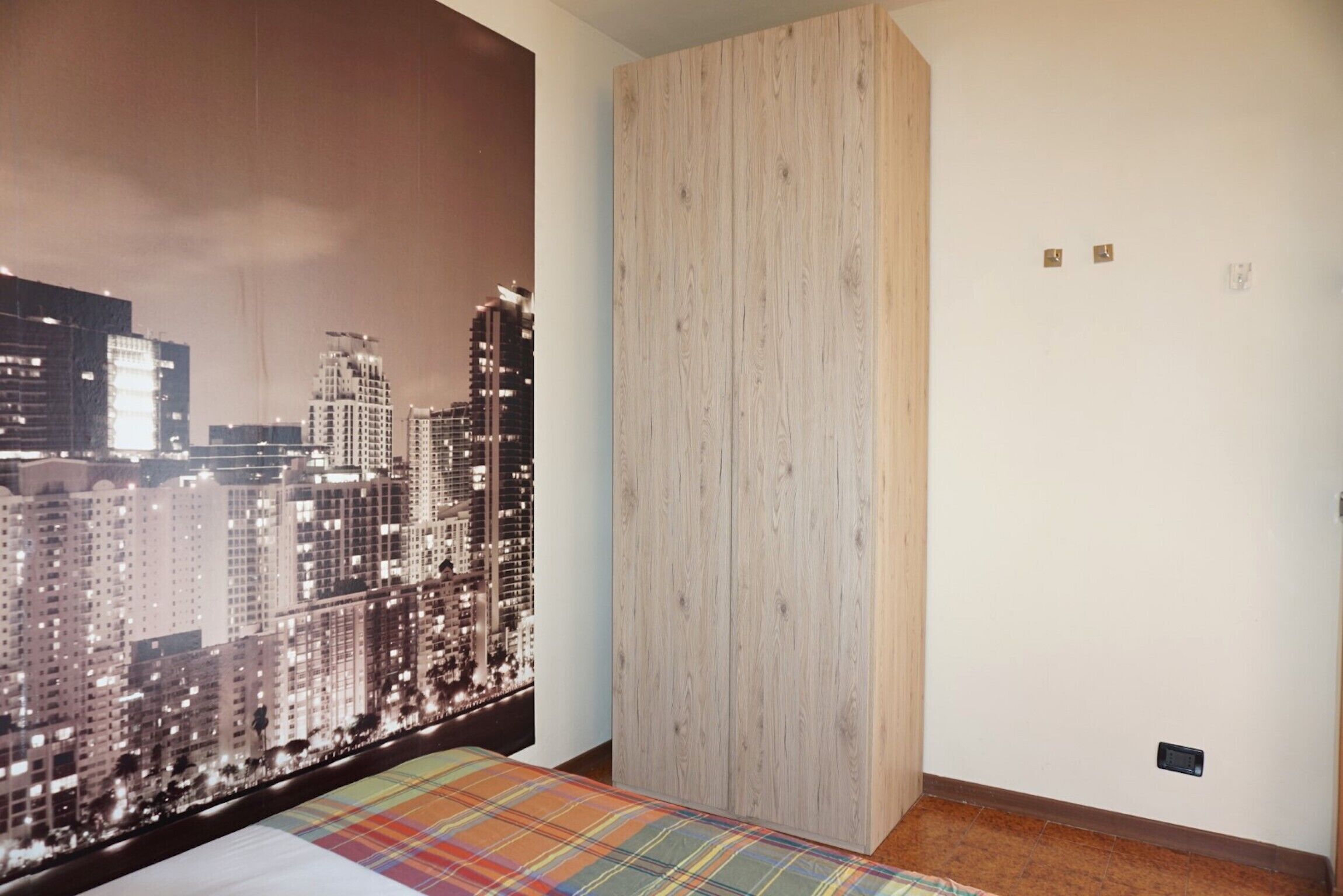 A bedroom with a cityscape view, a wooden wardrobe, and a plaid bedspread.