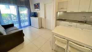 A modern living room with a sofa, a dining table with chairs, a kitchen area with cabinets and a sink, and a sliding glass door leading to a balcony. - Relax by the Garden-flat for up to 6 Guests (Caorle)