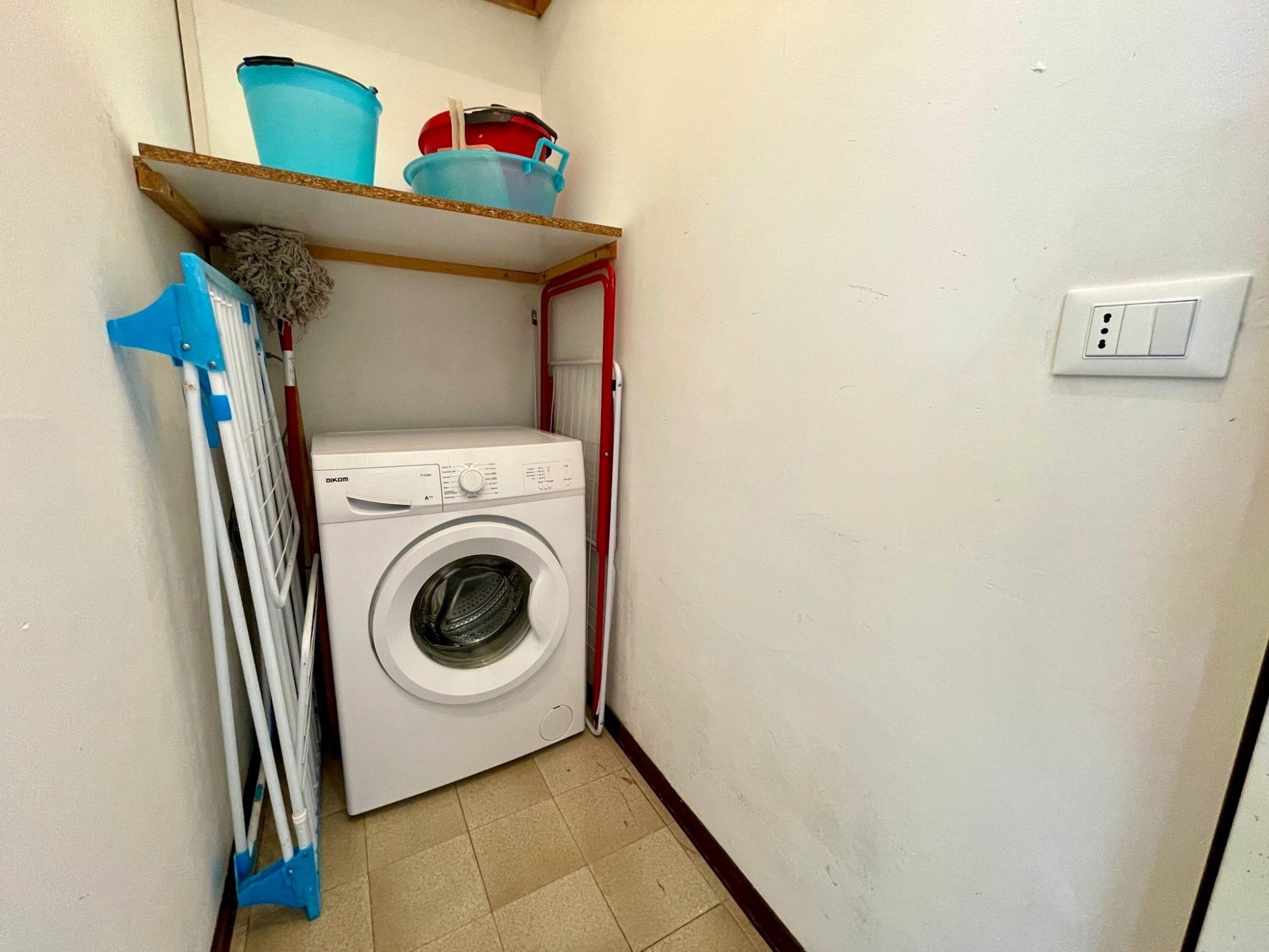 Uma lavanderia compacta com máquina de lavar, um varal e uma prateleira com recipientes de plástico.