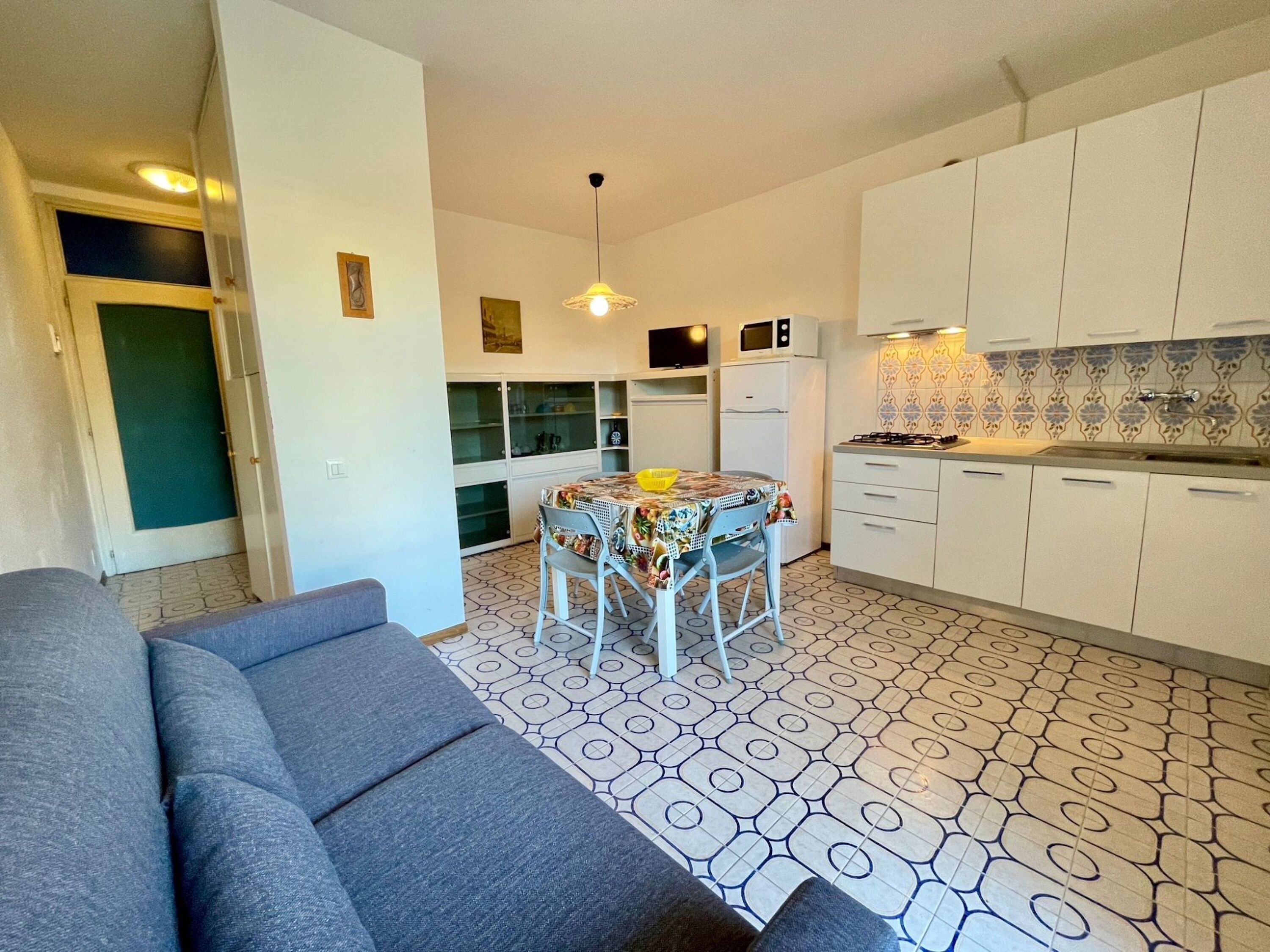 Un espai habitable compacte amb un sofà, taula de menjador amb cadires, petita cuina i paviment estampat.