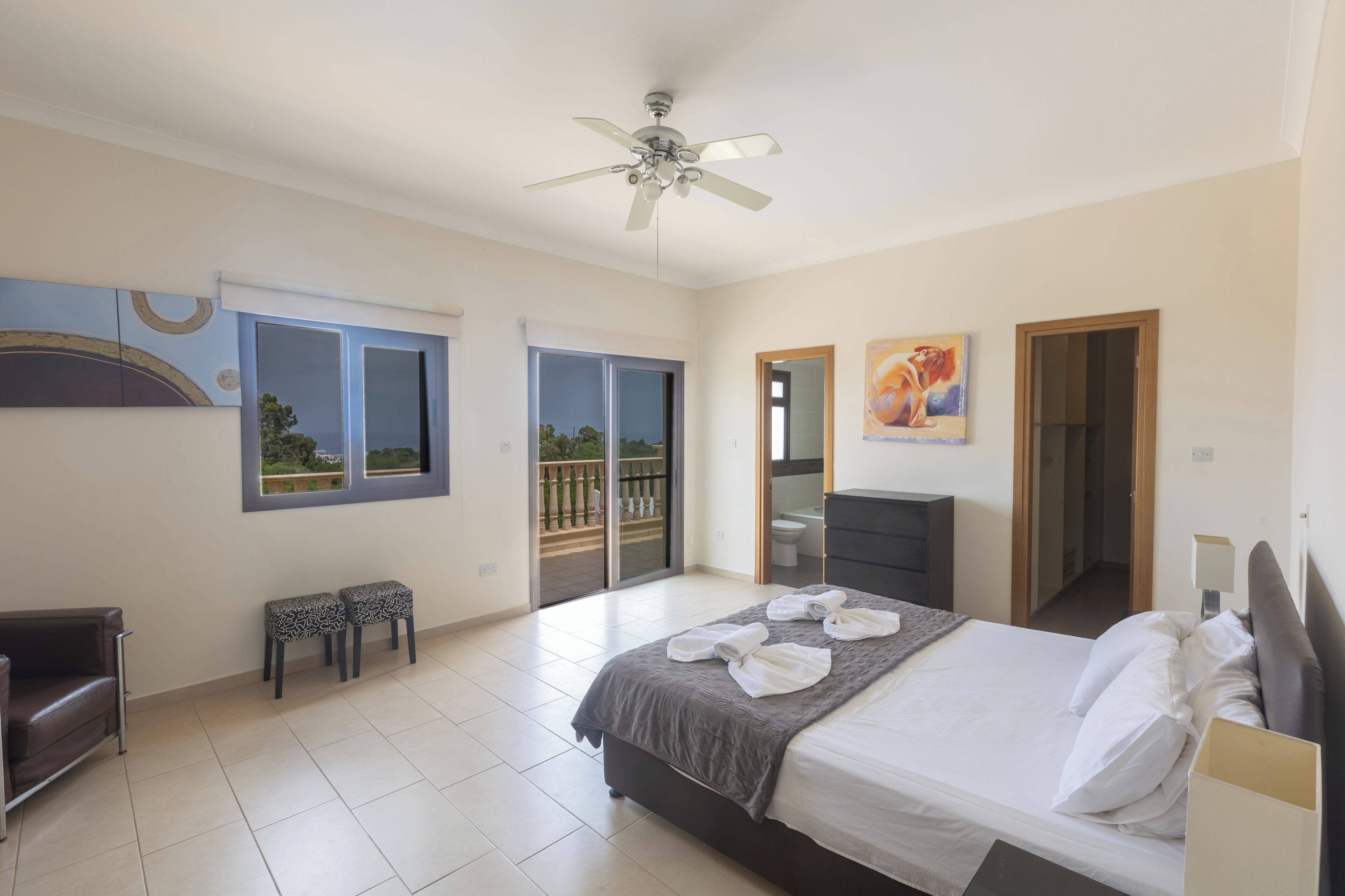 A hotel room with a bed, a ceiling fan, a painting, a desk, a chair, and a view of the outdoors.