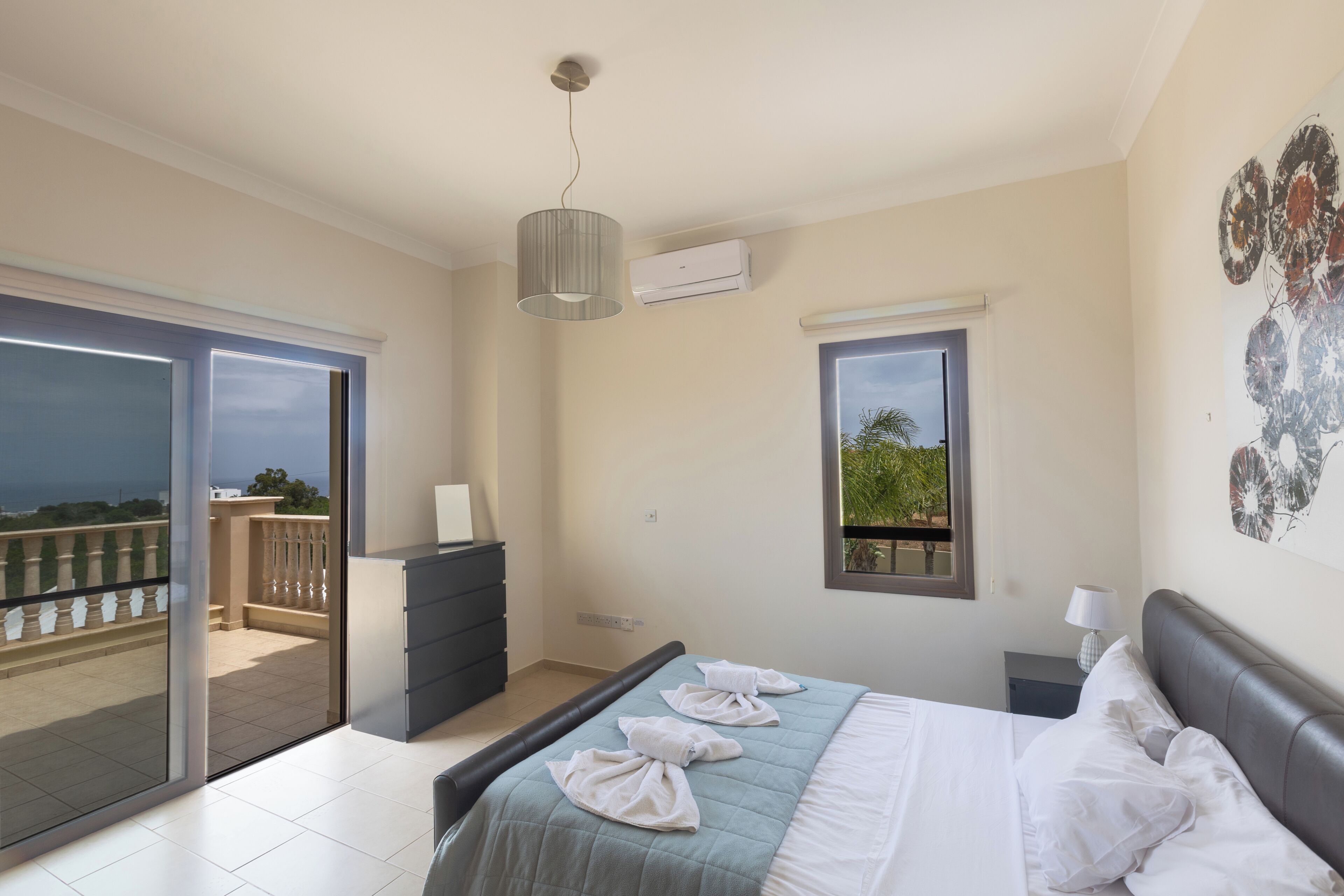 A bedroom with a bed, a dresser, a nightstand, a lamp, a window, and a balcony.