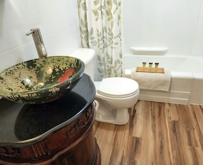 A bathroom with a wooden vanity, a green glass bowl sink, a white toilet, and a bathtub with a towel rack. - the Design Apartment at Sanctuary of the Swan Wellness Hotel Swan Rescue Farm (Birdsboro)