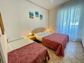 Hotellihuone, jossa on kaksi sänkyä, yöpöytä lampun kanssa, seinä, jossa on kolme kehystettyä kuvaa, ja ikkuna, jossa on ohuet verhot.