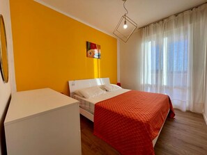 A hotel room with a bed, a white bedside table, a red bedspread, a painting on the wall, and a window with curtains. - Modern Apartment With Pool and sea View (Caorle)