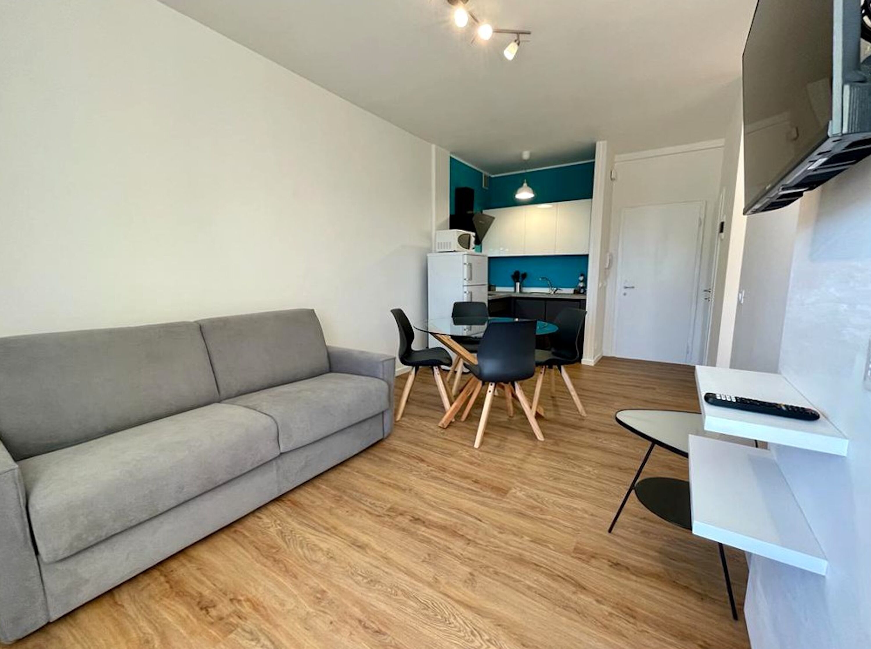 Een moderne woonkamer met een grijze bank, een eettafel met stoelen, en een keuken in de achtergrond.