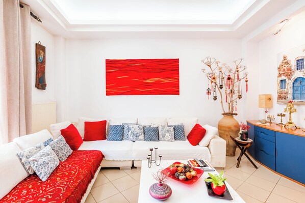 Ein modernes Wohnzimmer mit einem weißen Sofa, roten und blauen Kissen, einem roten abstrakten Gemälde, einer Vase mit Zweigen und einem kleinen Tisch mit Pflanze und Dekoration.