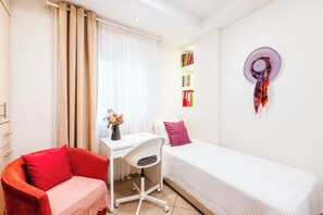 Ein kleiner, gut beleuchteter Raum mit Schreibtisch, Stuhl, Bett und Bücherregal.