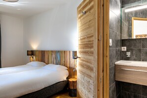 A bedroom with a wooden headboard, a bedside table, a nightstand, a bed, a wall-mounted light, and a bathroom with a sink and mirror. - Alps Retreat w/ Hot Tub (Abondance)