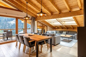 Et rummeligt værelse med træloft, et spisebord med stole, en sofa og et stort vindue med udsigt til et bjerglandskab.