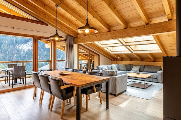 A spacious room with a wooden ceiling, a dining table with chairs, a sofa, and a large window offering a mountain view.