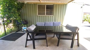 A patio area with a glass-top table and two cushioned chairs, a gas cylinder, and a covered seating area with a window.