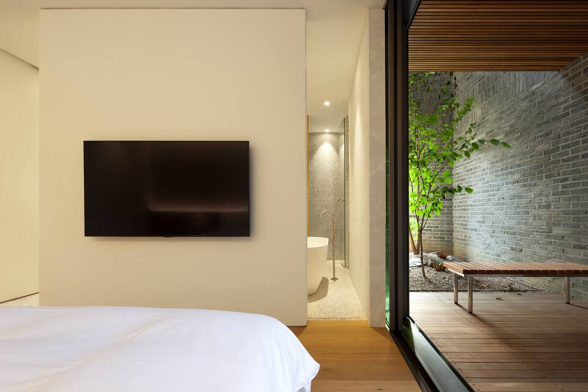 A modern bedroom with a large bed, a mounted TV, and a view of a brick wall and outdoor seating area.