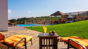 A poolside area with lounge chairs, a table with fruit and a drink, and a barbecue grill. - Luxury Endless Sea View Escape (Plaka)