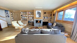 Un salon chaleureux avec un canapé gris, des étagères en bois, une cheminée et une grande fenêtre donnant sur l’extérieur.