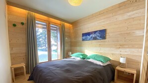 Une chambre à coucher aux murs lambrissés, avec un grand lit, deux tables de chevet, une petite table et une fenêtre avec des rideaux.
