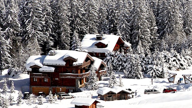 Un chalet enneigé, composé de plusieurs cabanes en bois, entouré d’une forêt dense.