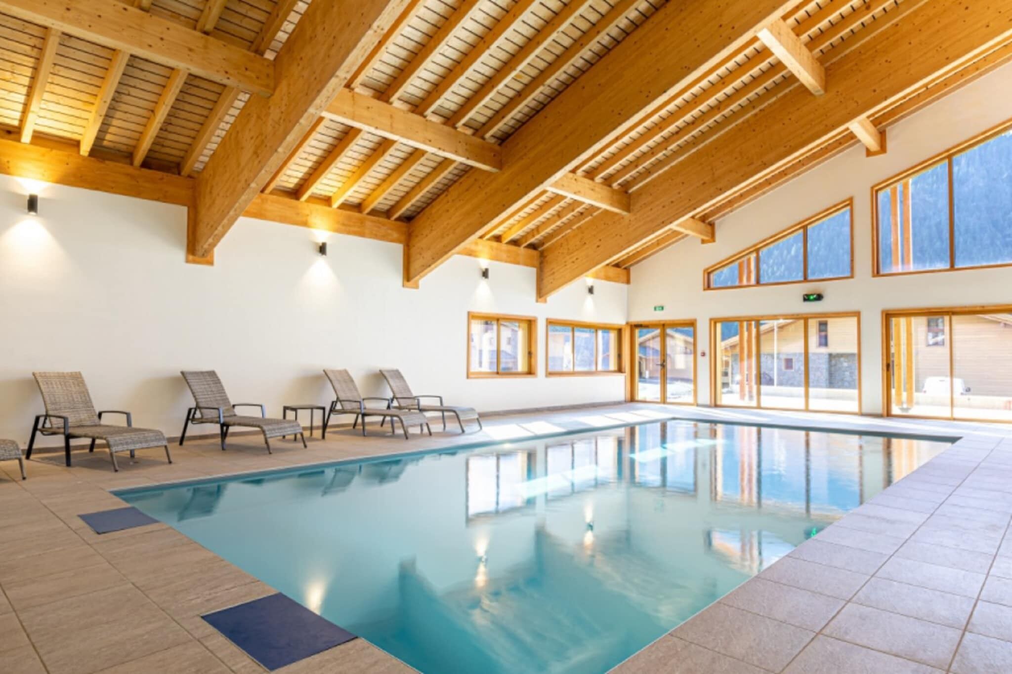Une piscine intérieure avec des poutres en bois, un sol carrelé et de grandes fenêtres laissant entrer la lumière du soleil.