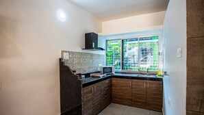 A kitchen with dark wood cabinets, a black countertop, a microwave, and a window with a view of greenery. - Whiteboard Homes (Siolim)