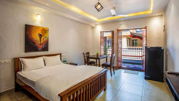 A bedroom with a bed, a table, chairs, a refrigerator, and a painting on the wall. - Whiteboard Homes (Siolim)