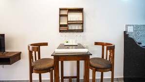 A dining area with a wooden table and chairs, a wall-mounted shelf with glasses, and a television on a shelf. - Whiteboard Homes (Siolim)