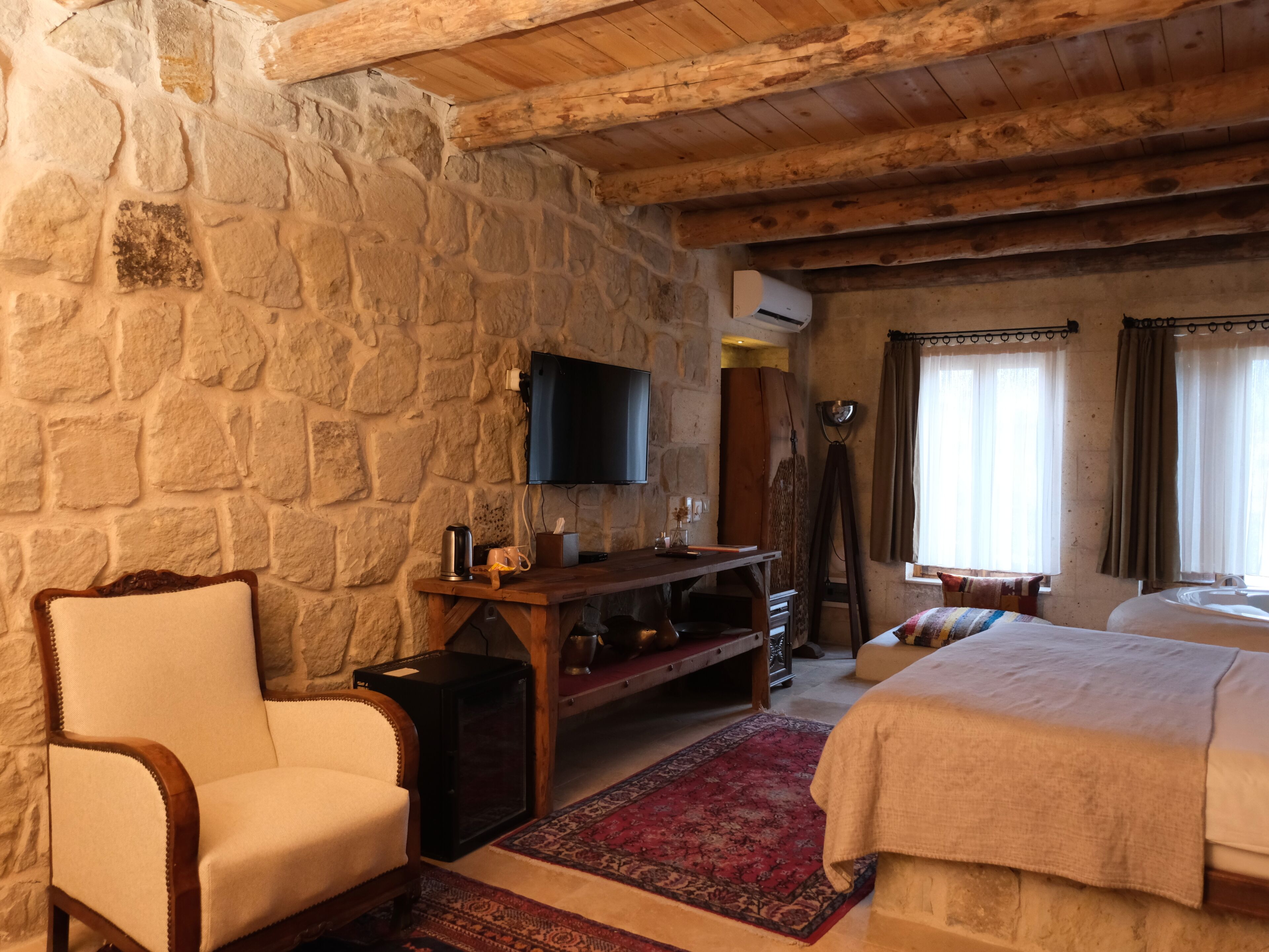 A stone-walled room with a bed, a chair, a TV, and a wooden ceiling.