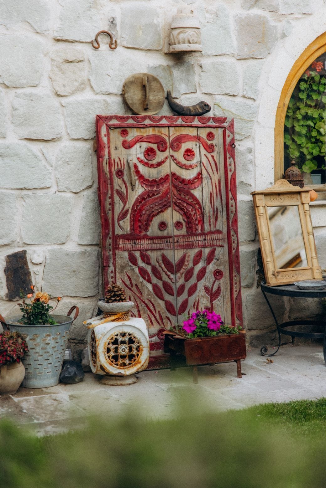 Un muro de piedra rústica con una puerta de madera decorativa, una maceta de metal con flores y un objeto de metal de estilo vintage.