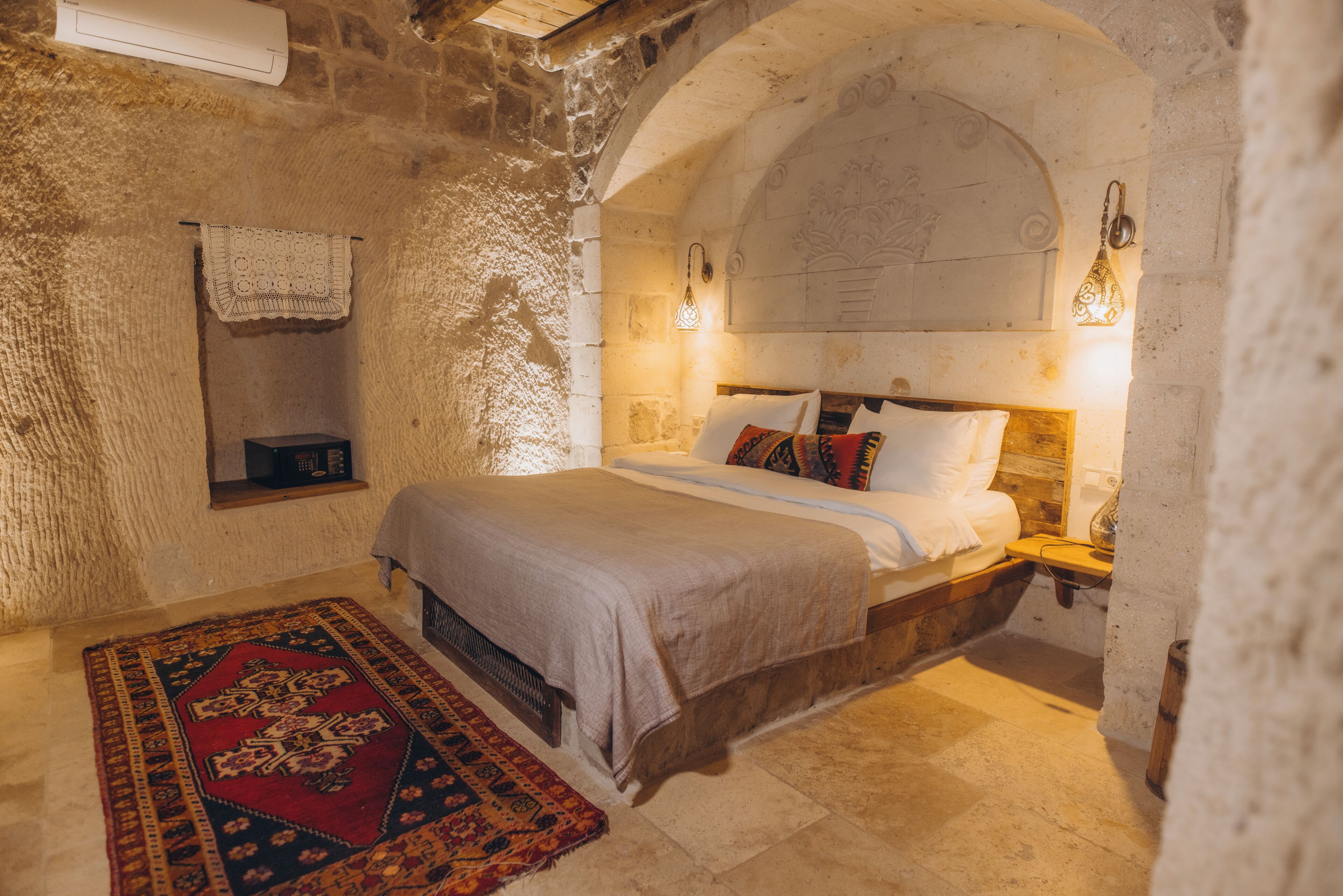 A stone-walled bedroom with a bed, a nightstand, a wall-mounted lamp, and a patterned rug.