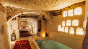 Una habitación con paredes de piedra, una bañera grande, una cama con un tapete rojo estampado y un ventanal circular.