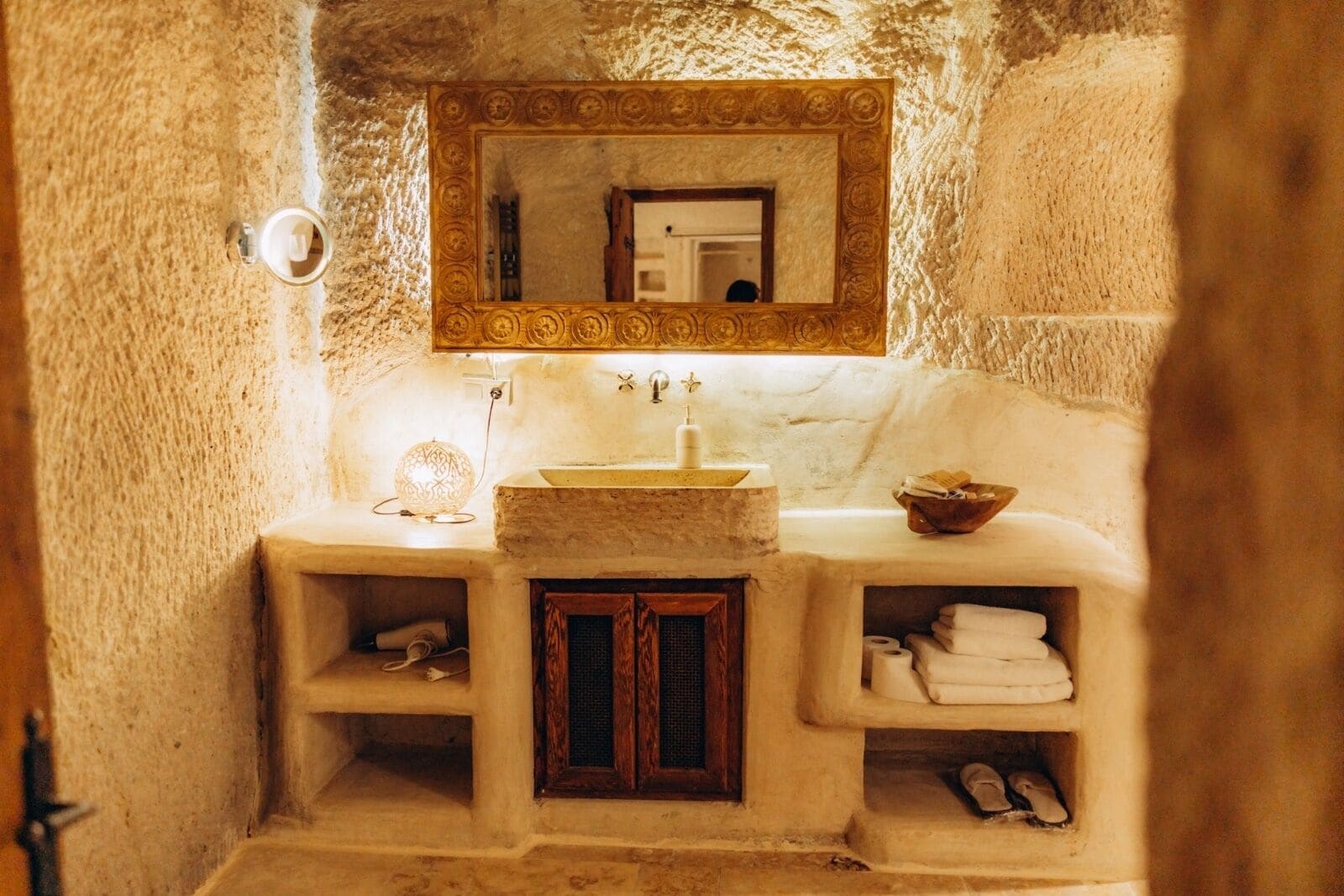 A bathroom with a stone sink, a mirror with an ornate frame, a lit lamp, and shelves with towels and toiletries.