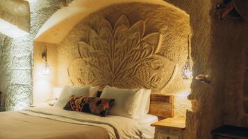 A stone-walled bedroom with a carved floral headboard, a wooden bedside table, and a bed with white linens and patterned pillows.