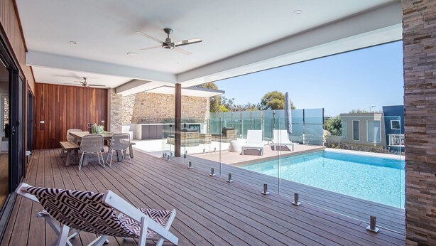Un área de piscina moderna al aire libre con terraza de madera, asientos y una sección con cerramiento de vidrio.