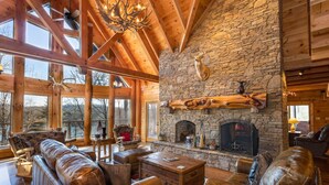 Un salón amplio con chimenea de piedra, mobiliario de cuero y ventanas grandes con vistas a un paisaje pintoresco.