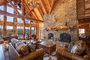 Un salón amplio con chimenea de piedra, mobiliario de cuero y ventanas grandes con vistas a un paisaje pintoresco.