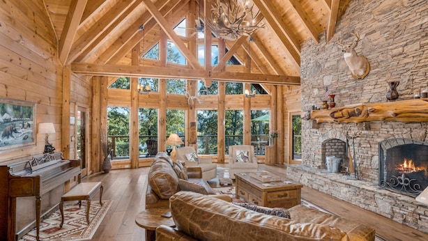 Un interior de cabaña de madera espaciosa con chimenea de piedra, muebles de cuero y ventanas amplias que ofrecen vistas al exterior.