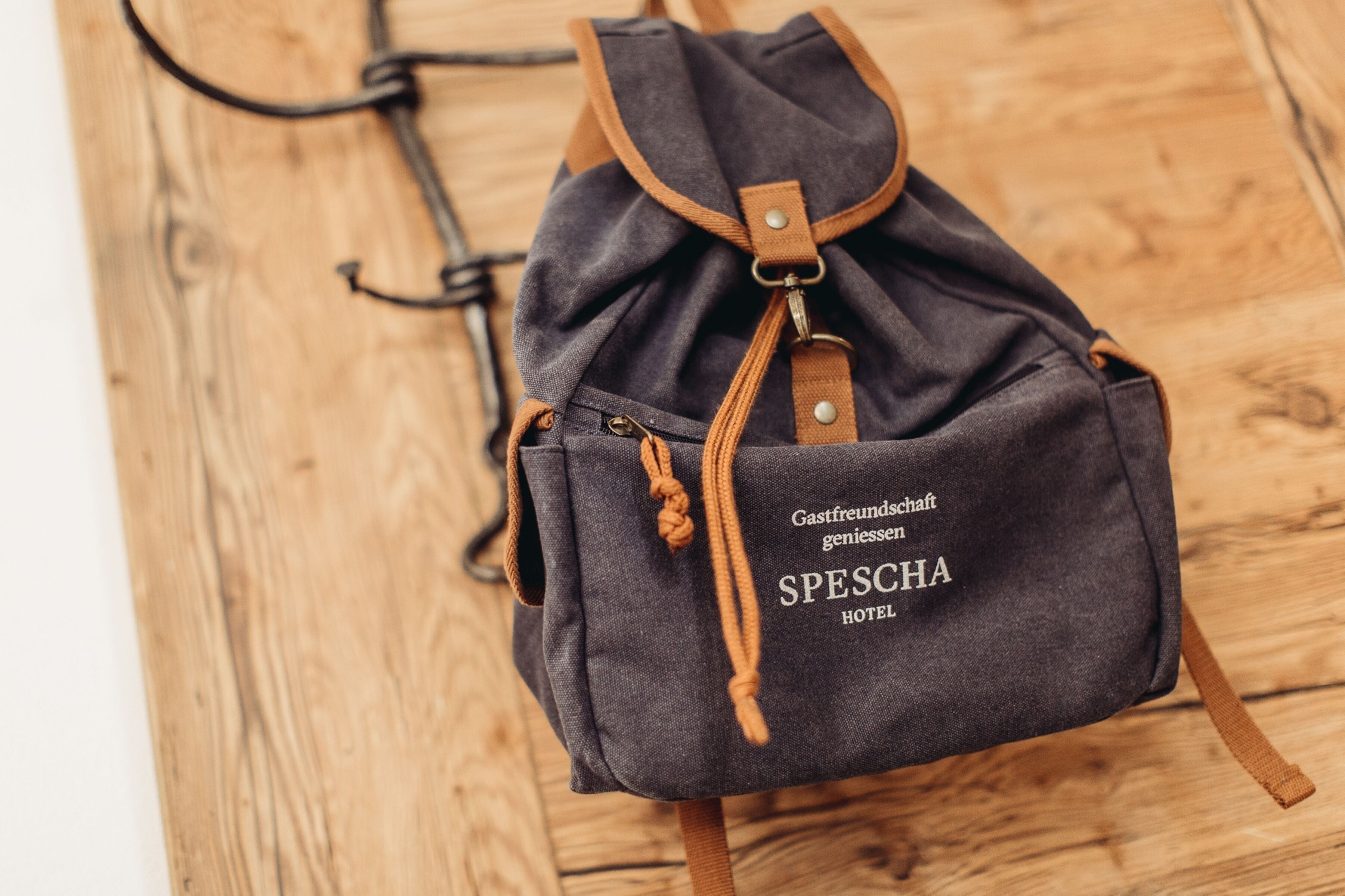 A dark gray drawstring bag with "Gästfreundschaft genießend SPESCHA HOTEL" printed on it, placed on a wooden floor.