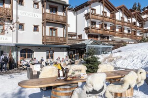 Un área de comedor exterior cubierta de nieve con mesas y sillas de madera, una mesa de buffet con comida y bebidas, y un edificio con balcones y el nombre "SPESCHA" a la vista.