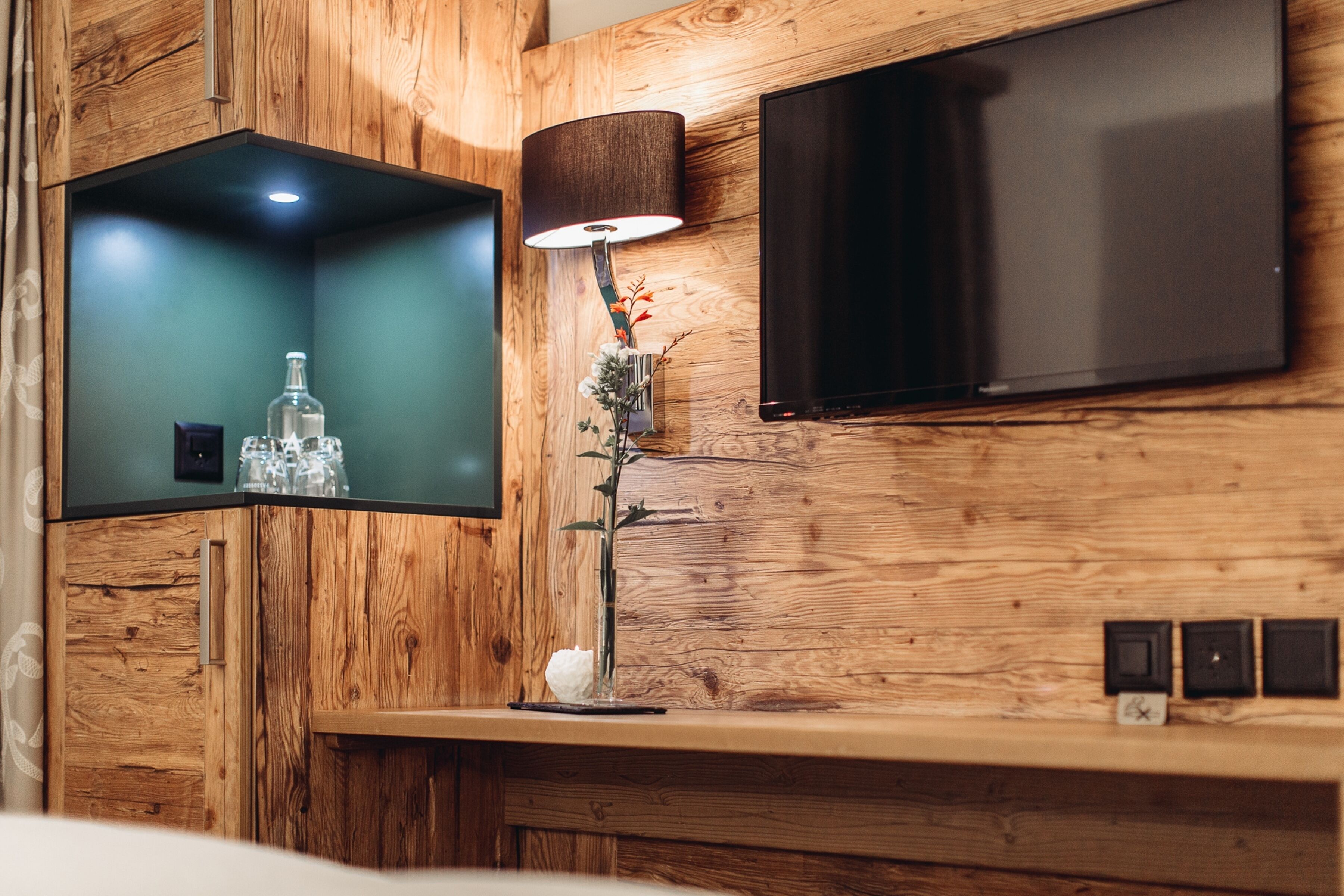 A modern room with a wooden wall panel, a flat-screen TV mounted above a wooden shelf, and a table lamp with a vase of flowers.