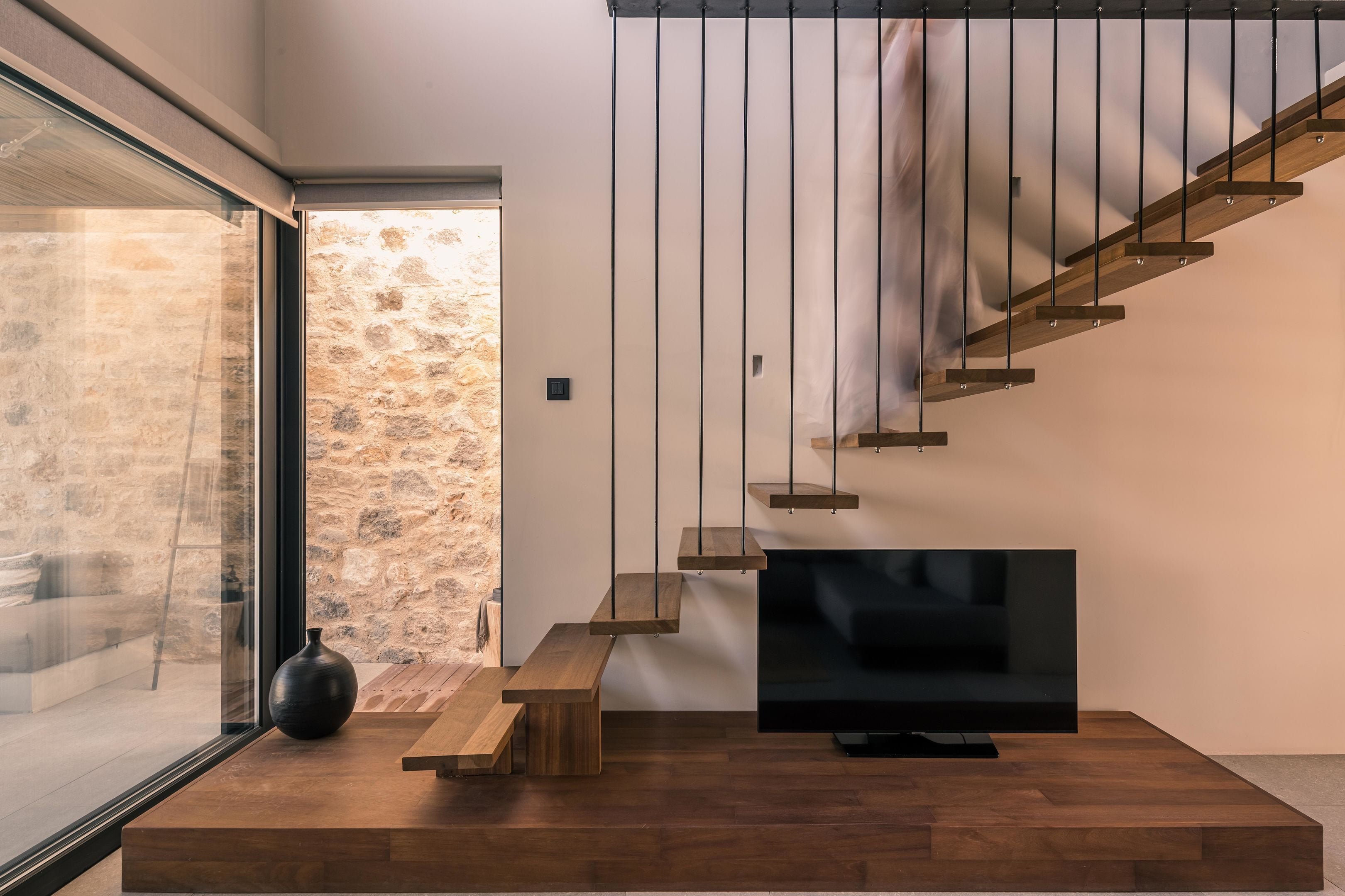 A modern living room with a wooden staircase, a flat-screen TV, and a stone wall.