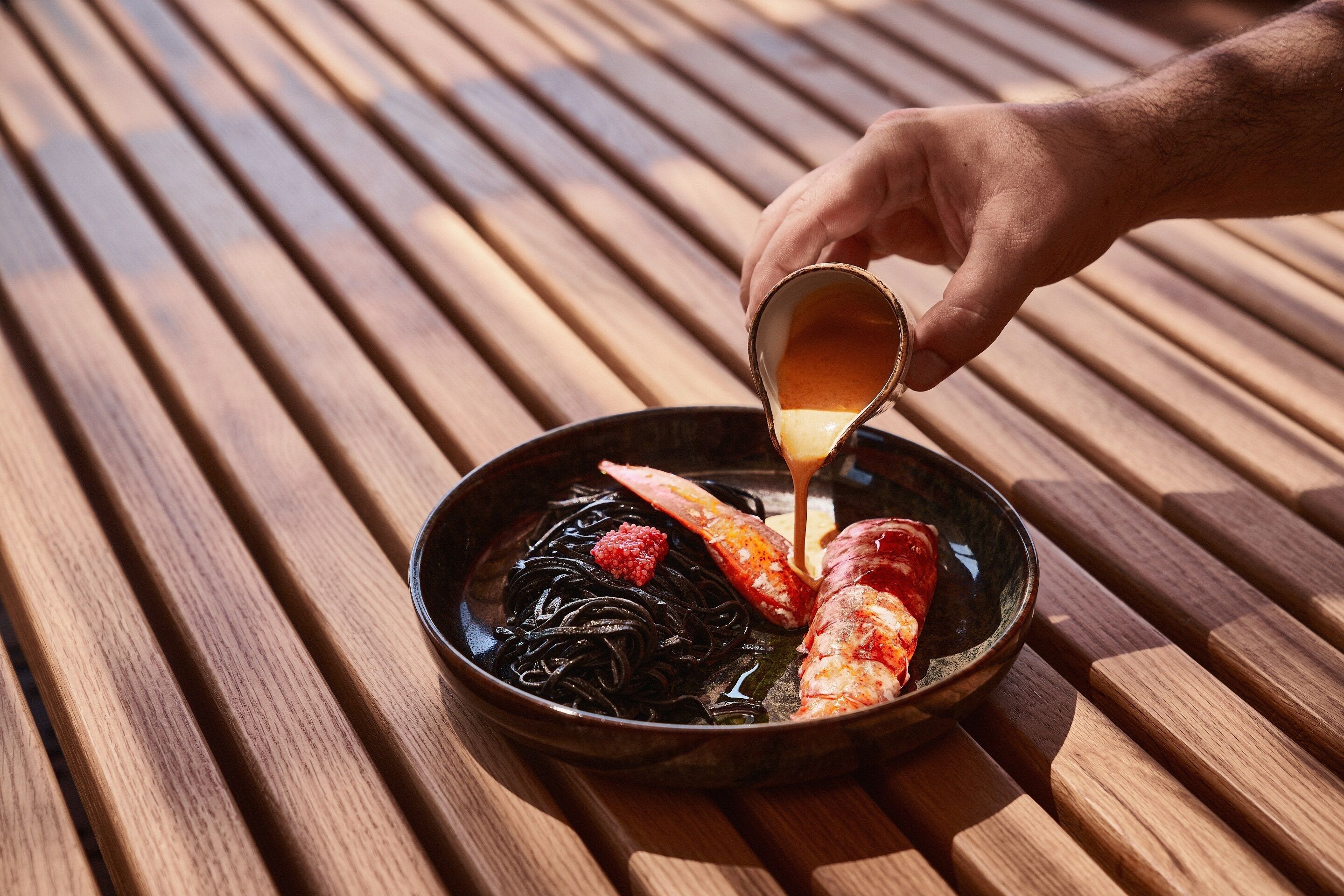 Una mano sirviendo salsa sobre un plato de mariscos y fideos sobre una mesa de madera.